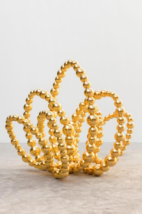 Une sculpture contemporaine composée de sphères dorées brillantes, reliées entre elles pour former des boucles sinueuses et aériennes. Chaque boucle rappelle la forme d'un pétale, créant ensemble une structure florale abstraite qui évoque un lotus stylisé. L’œuvre est posée sur un sol en béton clair, dans un espace minimaliste aux murs blancs, mettant en valeur l’éclat doré des sphères.