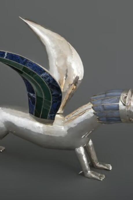 Sculpture en métal poli représentant une créature mythologique hybride, dotée d’un corps de lion et d’ailes incrustées de motifs colorés. L’œuvre se distingue par ses lignes stylisées et son expression saisissante.