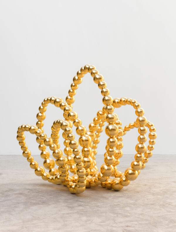Une sculpture contemporaine composée de sphères dorées brillantes, reliées entre elles pour former des boucles sinueuses et aériennes. Chaque boucle rappelle la forme d'un pétale, créant ensemble une structure florale abstraite qui évoque un lotus stylisé. L’œuvre est posée sur un sol en béton clair, dans un espace minimaliste aux murs blancs, mettant en valeur l’éclat doré des sphères.