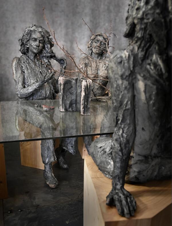 Détail de l'œuvre "À la table des Dames" de Cécile Raynal, où figure une table centrale en verre, et autour trois femmes sculptées en grès. Deux nous font face, une est de dos, posées toutes trois sur des socles en bois. Au centre, l'évocation troublante d'une nature avec des branchages et une paire de ciseaux...