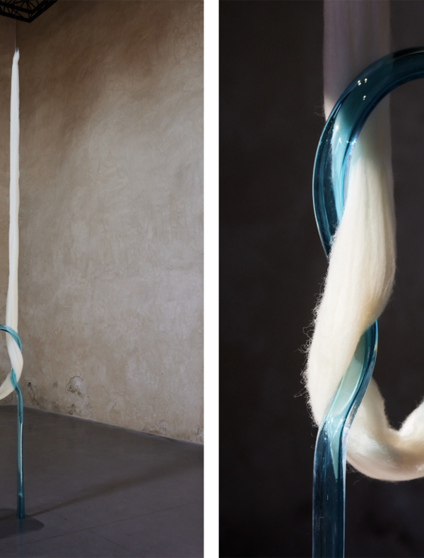 Une installation artistique contemporaine montrant une structure en verre bleu transparent en forme de boucle verticale. Deux longues mèches de fibres blanches (ressemblant à de la laine ou du coton) passent à travers la boucle et tombent jusqu'au sol. L’image de gauche montre l’installation entière dans une pièce aux murs en plâtre beige, tandis que l’image de droite offre un gros plan sur la texture des fibres et la transparence du verre.