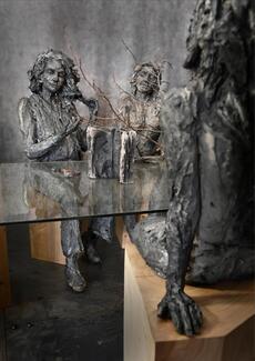 Détail de l'œuvre "À la table des Dames" de Cécile Raynal, où figure une table centrale en verre, et autour trois femmes sculptées en grès. Deux nous font face, une est de dos, posées toutes trois sur des socles en bois. Au centre, l'évocation troublante d'une nature avec des branchages et une paire de ciseaux...
