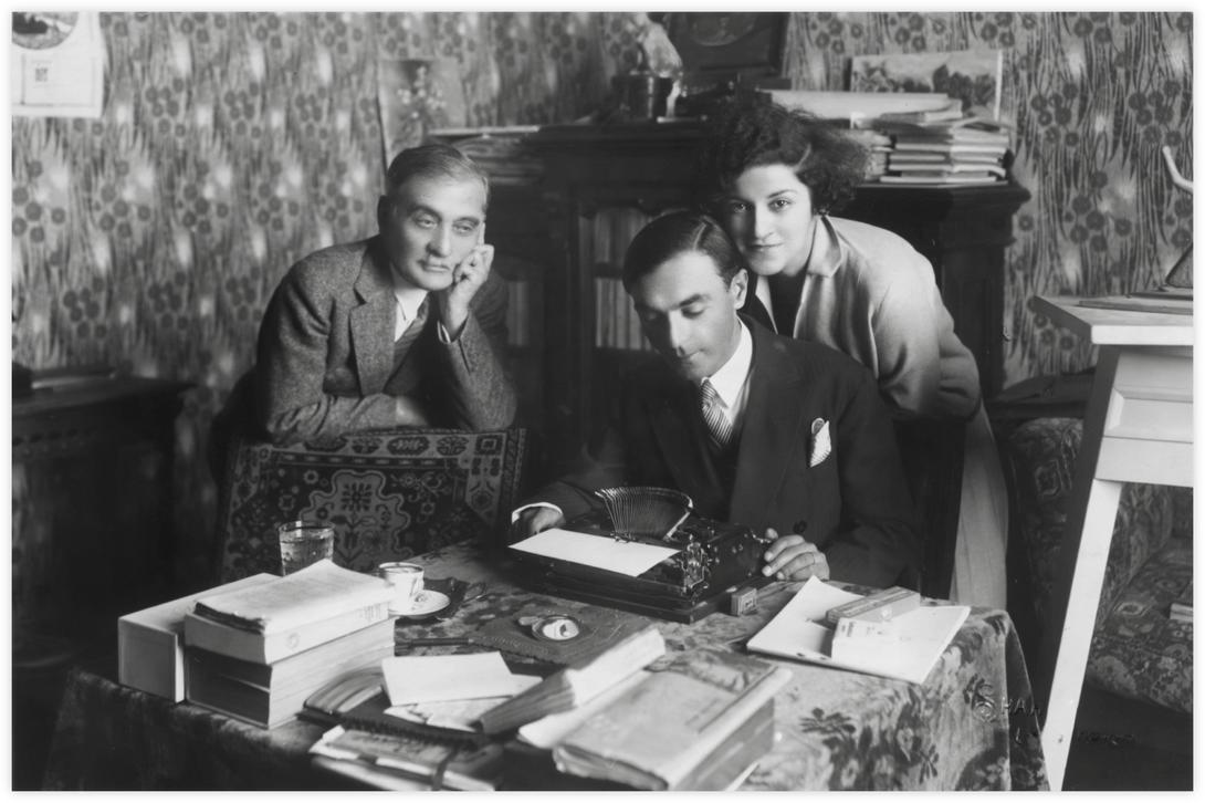 24 Daria, Imre et Armenag, Alexandrie, Photo Shahen vers 1930 Daria se penche tendrement sur le dos d'Imre, qui reste concentré sur la feuille blanche dans sa machine à écrire. Armenag, le père de Daria se joint à eux. Il y a une tasse de café et un verre d'eau posés devant lui. Il s'appuie sur un fauteuil tapissé d'un beau tapis. La table est aussi recouverte d'un tapis épais et plusieurs livres et journaux sont posée devant.