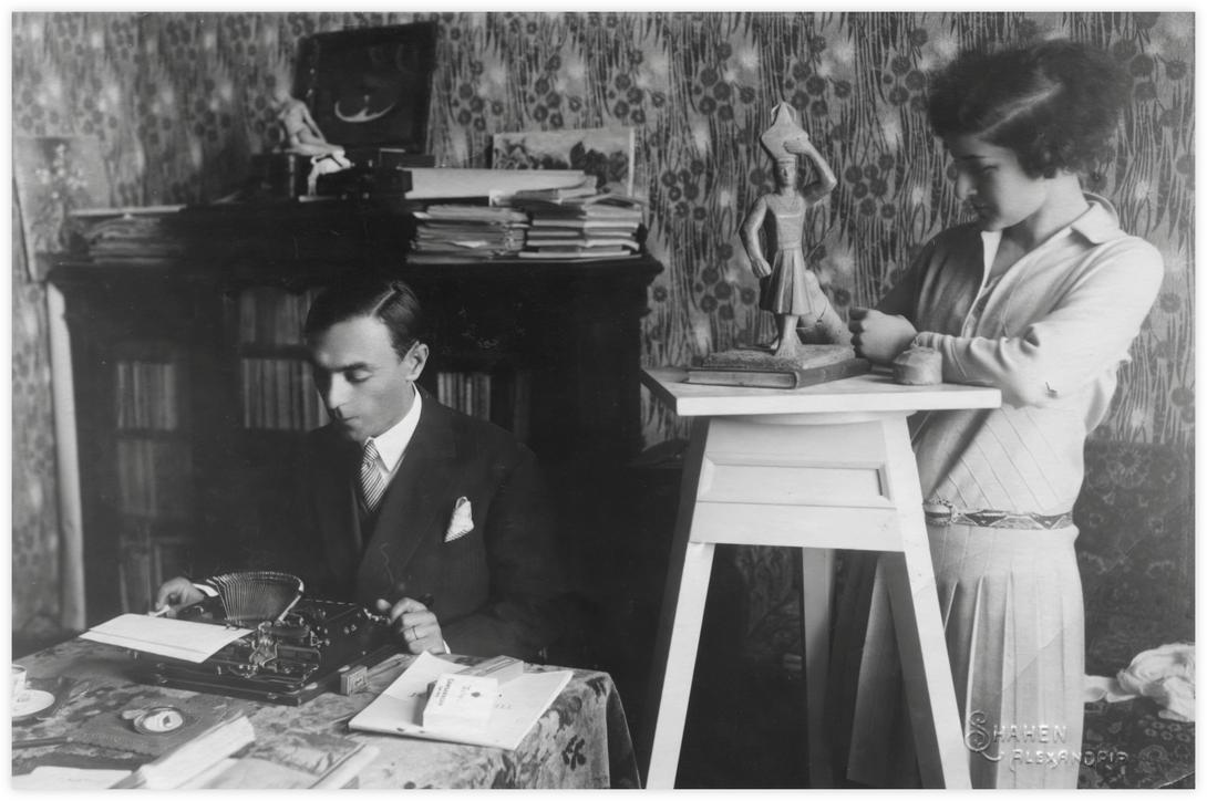 25 Daria et Imre Alexandrie, Photo Shahen vers 1930. Daria et Imre posent dans la maison familiale de Daria à Alexandrie. On voit un papier peint imprimé typique du style de intérieurs anglais de l’époque. Daria examine une sculpture de "la porteuse d'eau" posée sur une stèle. Imre est concentré devant sa machine à écrire. Il y a un paquet de cigarettes Gamsaragan posé sur la table.